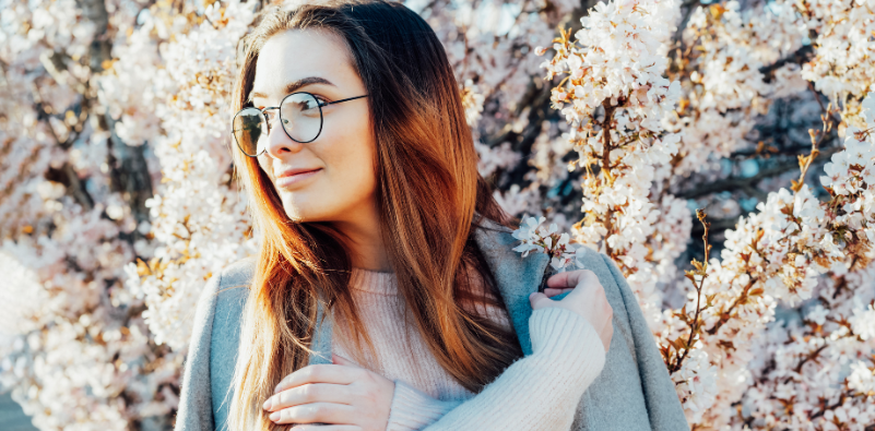 A young woman outside next to a tree with white blossoms, wearing round glasses.