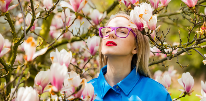 A young woman wearing a pair of pink glasses outside next to a tree with pink and white flowers.