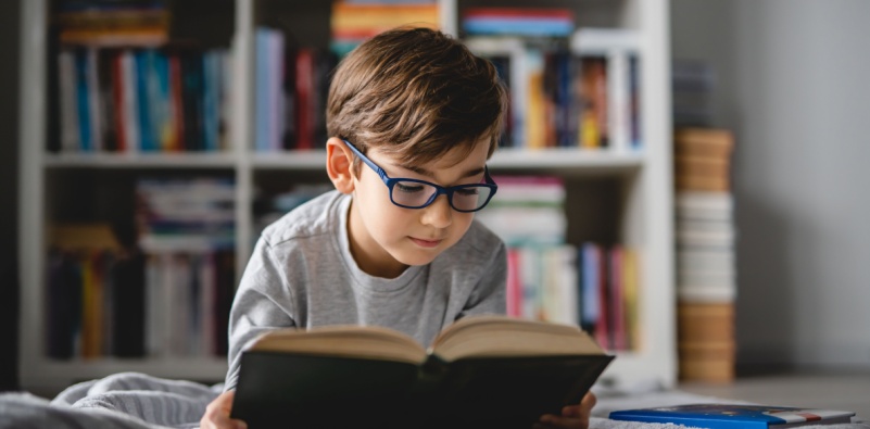 A young boy wearing a pair of glasses reading a book in his bedroom.