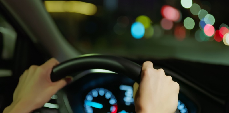 A close-up of a woman’s hands on the steering wheel, driving at night, with blurred streetlights in the background