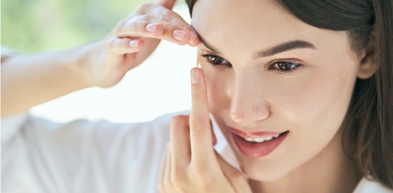 Woman with dark brown hair holding a contact lens on her finger ready to insert into her eye.