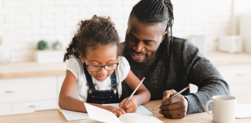 Alt text – A dad helping his young daughter, who is wearing glasses, do homework in the kitchen.